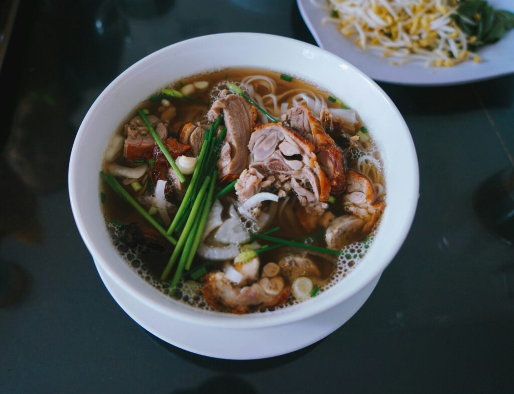 A white bowl of duck noodle soup with flat rice noodles, roasted duck pieces, green scallions, and white onions, served on a dark table with a blurred plate of bean sprouts in the background.